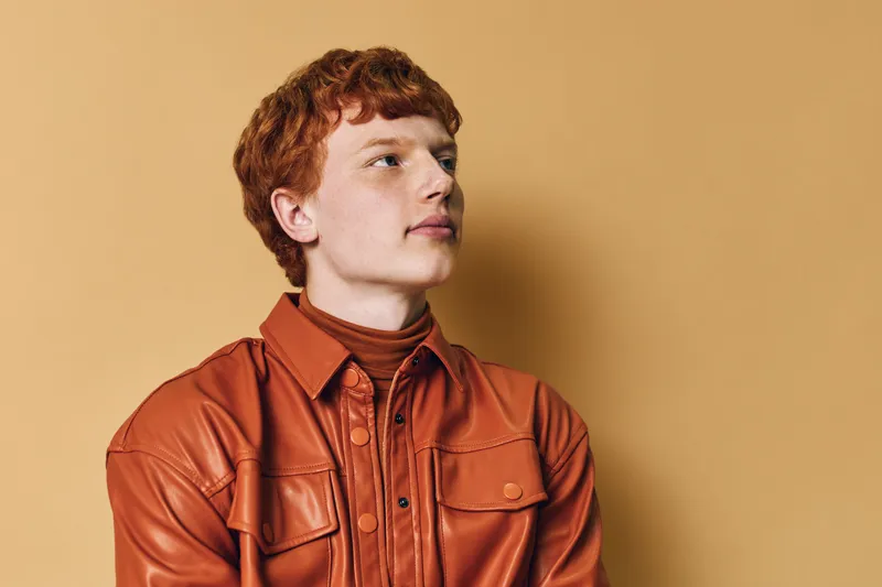 Young man with red hair wearing brown leather jacket and turtleneck posing against beige background for fashion portrait and style concept in studio.