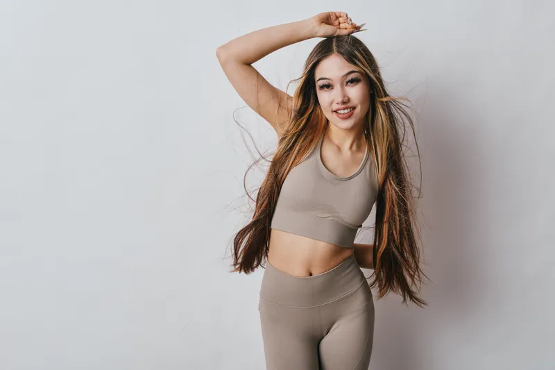 Young woman with long hair in beige activewear posing with hand over head smiling for fitness, health and lifestyle on white background in studio space.