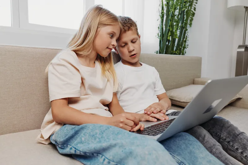 Two children sitting on couch using laptop together, focused on screen in bright living room. Concept of learning, technology, and collaboration between kids.
