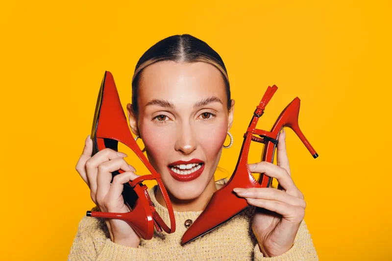Young woman with slicked hair holds red high heels near face on bright yellow background. Concept of fashion, style, footwear, and vibrant color contrast in studio portrait.
