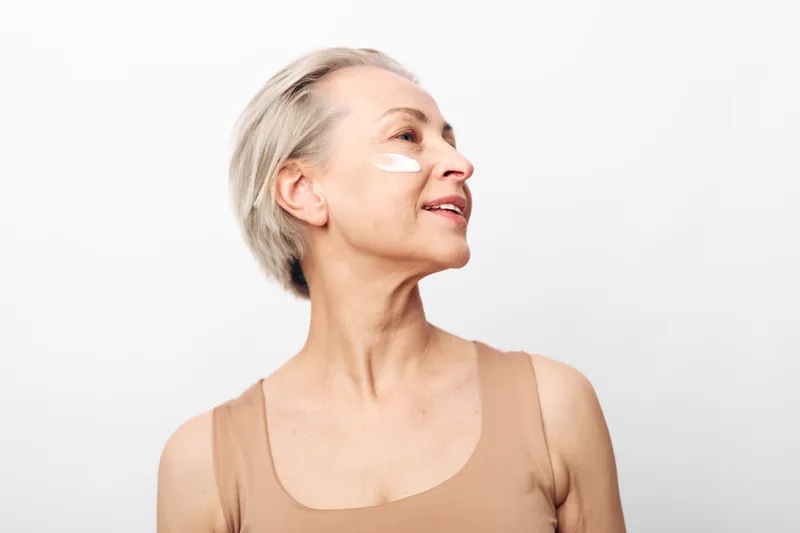 Smiling mature woman with short blond hair applies face cream on cheek. Skincare concept for aging skin, self-care, natural beauty, and healthy lifestyle on white background.