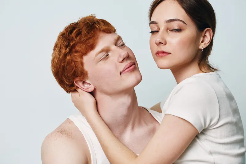 Couple portrait with young man and woman embracing, closeup with soft expressions and simple casual clothing on light neutral background in studio.