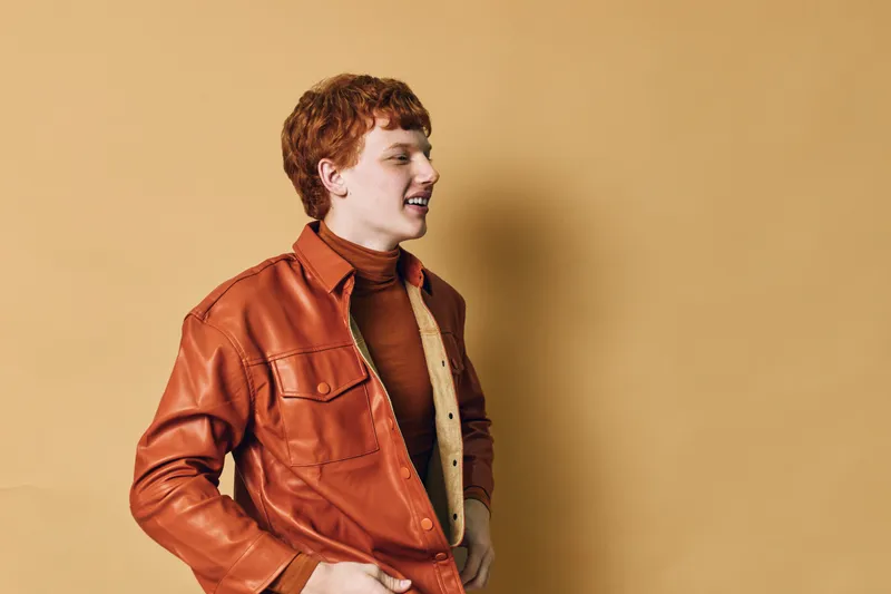 Young man with red hair wearing a brown leather jacket and orange turtleneck smiling in front of beige background. Casual style with confident and happy expression in studio.