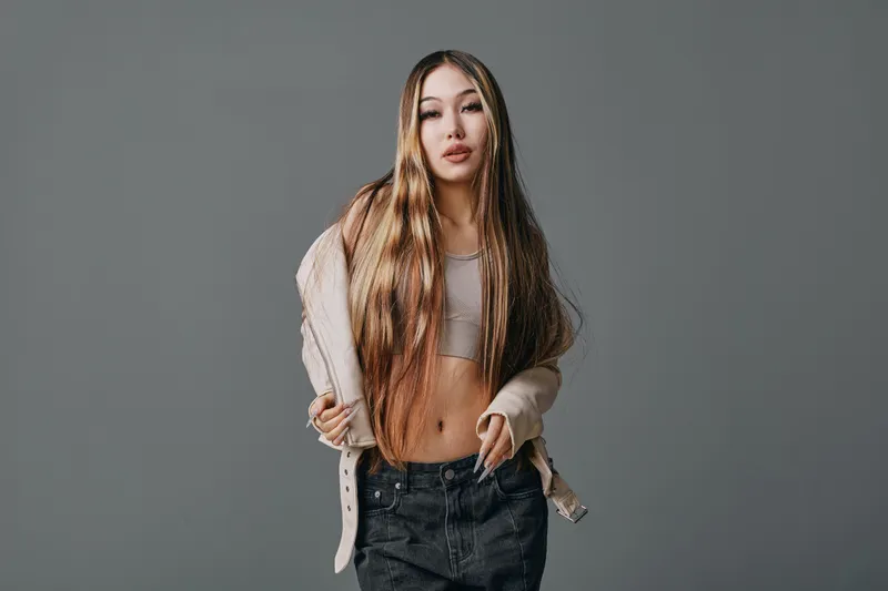 Young woman with long straight hair wearing casual beige sweater and crop top posing confidently against grey background in studio. Fashion, beauty and style.