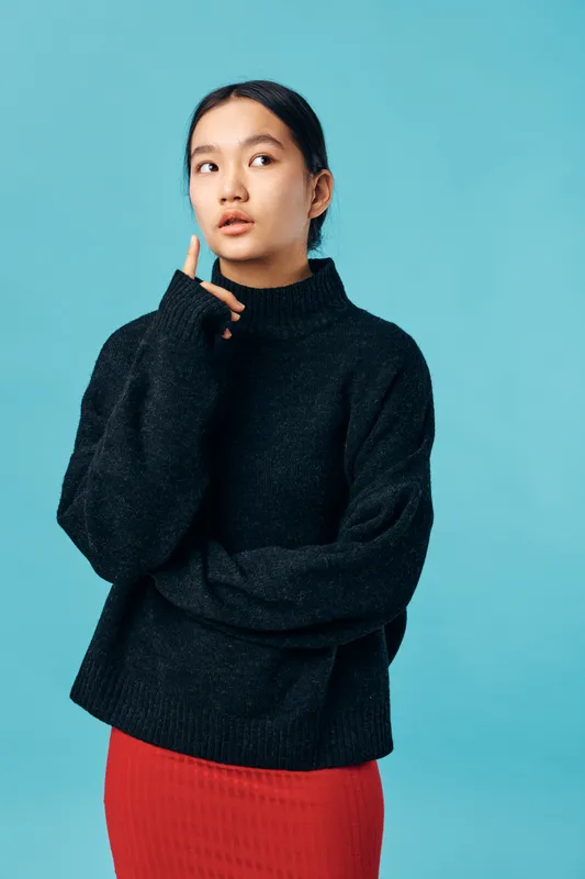 Young woman in black sweater and red skirt stands against blue background looking sideways with thoughtful expression and hand near face. Casual fashion portrait with studio lighting.