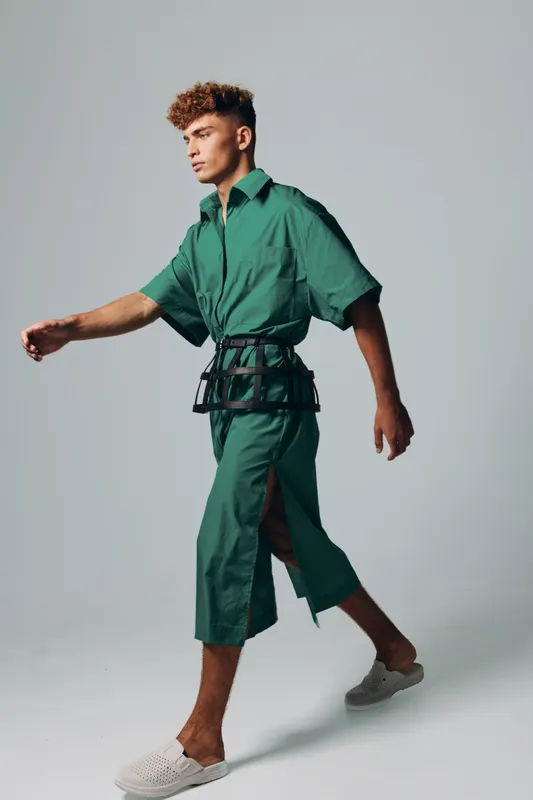 Young man walking in green oversized shirt and matching wide-leg pants with side slit, wearing white slip-on shoes and black belt accessory on plain background in studio.