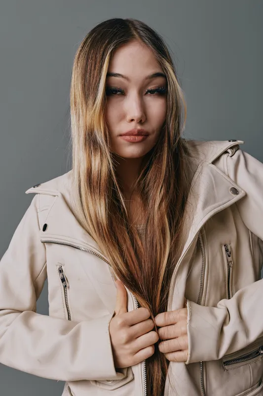 Young woman with long straight hair wearing beige jacket holding collar and looking at camera with neutral expression against neutral background studio portrait.