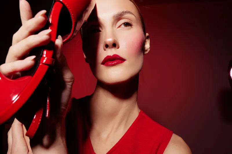 Close-up of a woman with red lipstick and red dress holding a red high heel shoe near her face. Studio lighting highlights her smooth skin and elegant makeup on red background.