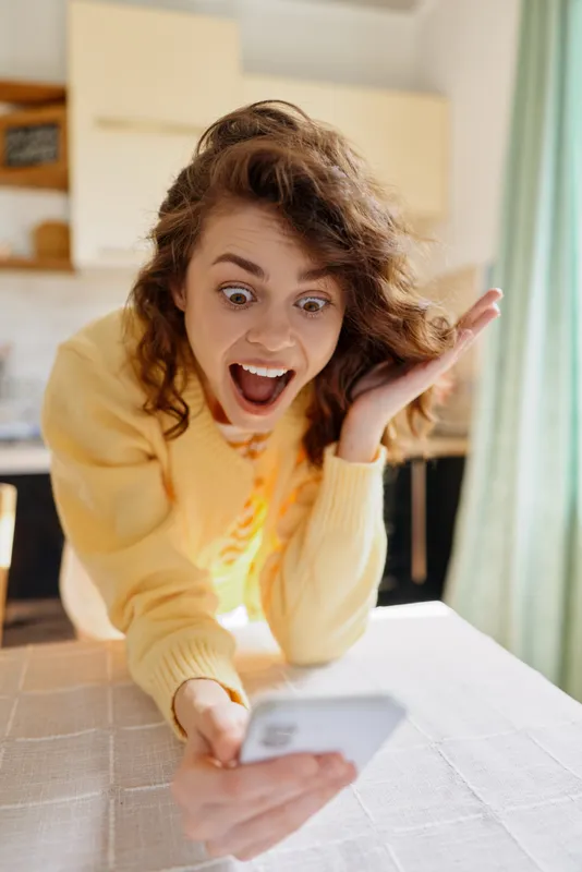 Woman at home reacts with surprise while looking at smartphone, wearing a cozy sweater, in a bright kitchen, an authentic candid moment of everyday communication