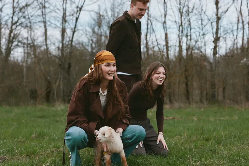 Three friends in a park share a light moment with a small dog, wearing casual autumn clothing. Natural setting, candid lifestyle, outdoor joy, companionship, and relaxed mood.