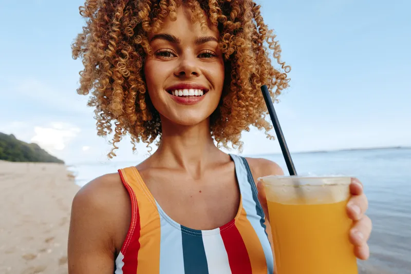 Happy woman at the beach wearing a striped swimsuit, holding a glass of orange juice, sunny day, smiling with a relaxed vacation mood, shoreline in the background