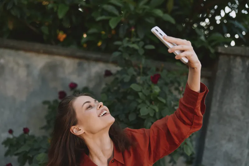 A smiling woman in a red sweater takes a selfie outdoors, lifting her phone above her head. A candid portrait capturing casual lifestyle and confident self expression in daylight