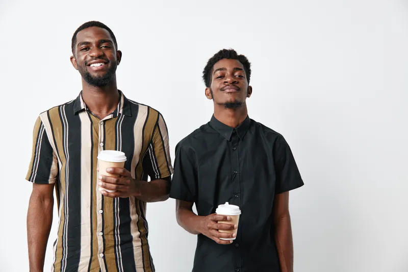 Two young African American men with dark skin holding coffee cups smiling and standing against white background in casual clothing for friendship and lifestyle.