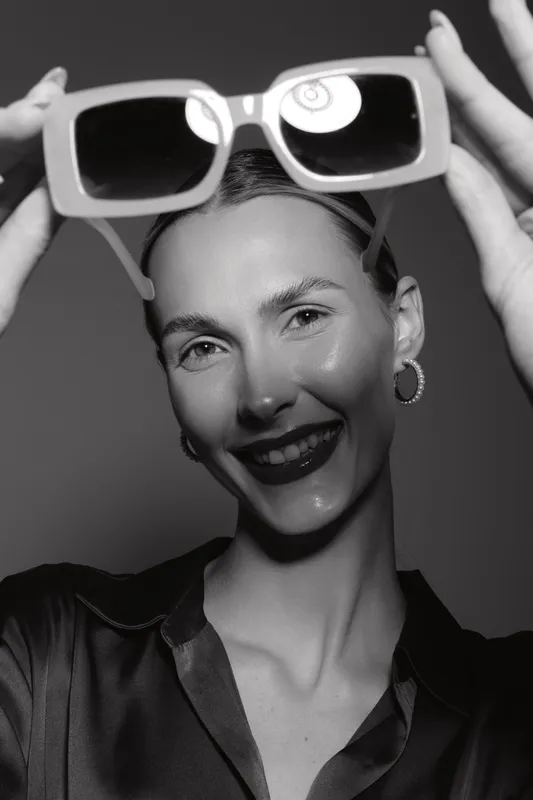 Young white woman with pale skin smiles holding sunglasses above her head in black blouse. Studio portrait with soft lighting. Concept of fashion, beauty, and eyewear accessory.