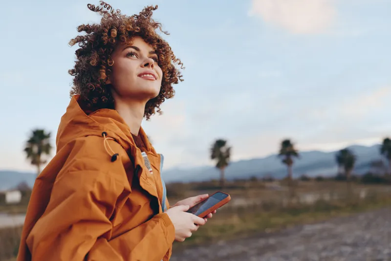 Woman with curly hair outdoors in an orange jacket, holding a smartphone and looking up at sunset sky. Travel lifestyle on a windy road, with nature landscape behind, soft warm light today.