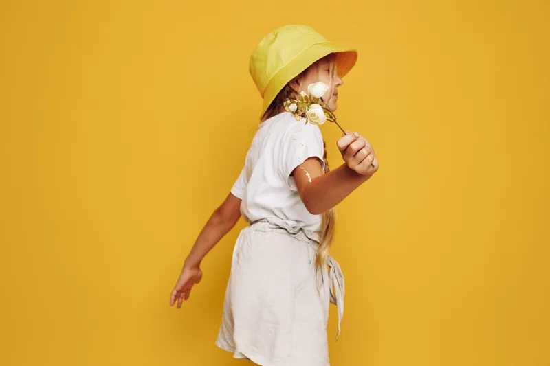 Little girl in yellow hat holding flowers spinning on yellow background. Casual outfit for spring or summer season.