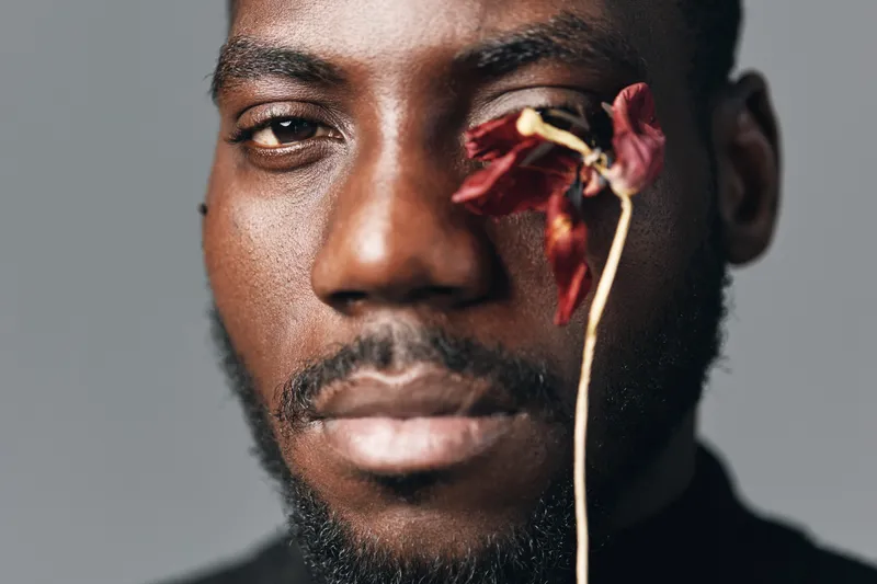 African man with dark skin and beard in creative conceptual style holding dried red flower near face on gray background. Closeup portrait with artistic expression and visual contrast.