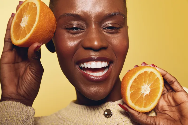 Happy African woman with dark skin holds two halves of fresh orange near face smiling with teeth on yellow background. Concept of healthy lifestyle, vitamin, skincare and wellness.