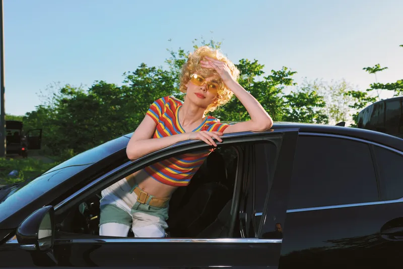 Smiling woman in striped crop top leaning from a car window on a sunny outdoor scene. Retro lifestyle and summer vibe with warm film tones and casual fashion.