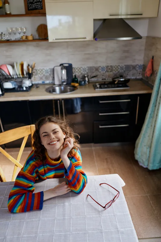 Young woman with curly hair smiles at the camera while resting her chin on her hand at a bright kitchen table, creating a warm, intimate home moment in a cozy setting.
