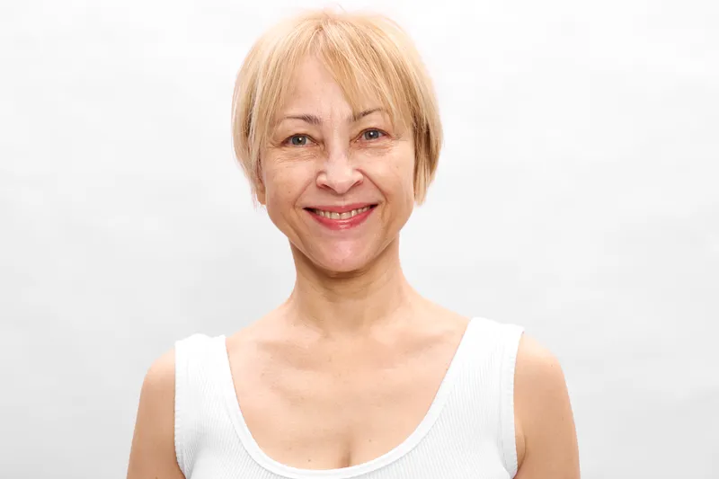 Smiling woman with short blonde hair posing portrait in a clean studio, wearing a white sleeveless top, suggesting skincare and wellness, soft light on face
