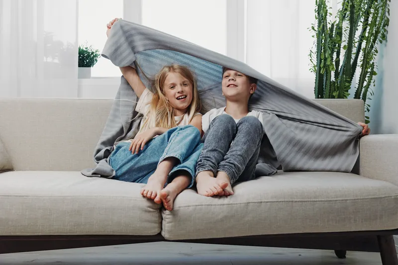 Happy children sitting on sofa under blanket smiling and playing together in bright living room interior with plants.