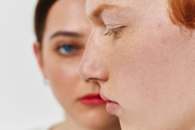 Closeup portrait of two women with freckles and natural skin, one in foreground with red hair and another blurred behind with blue eyes and red lips, studio lighting