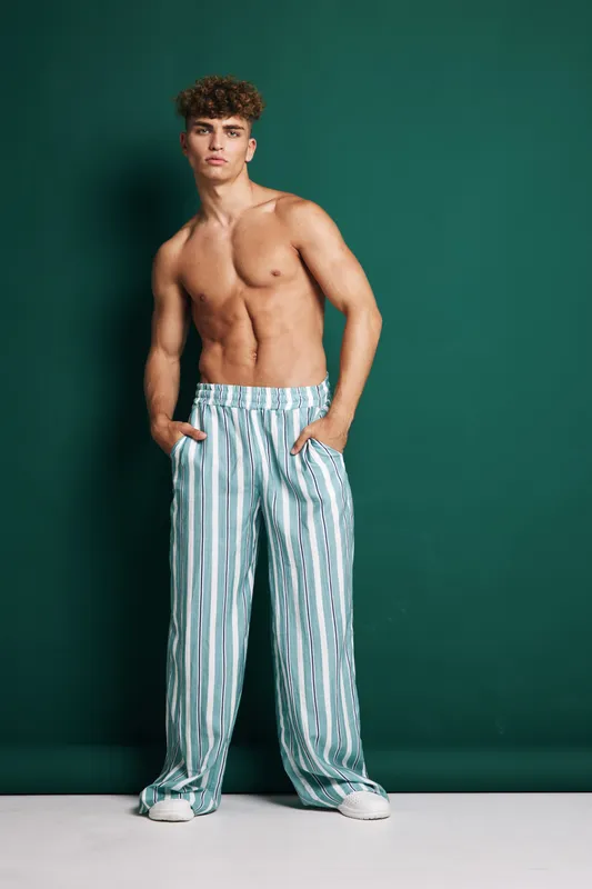 Young man with curly hair posing shirtless in striped wide pants and white sneakers against green background. Fashion model standing with hands in pockets and confident expression.