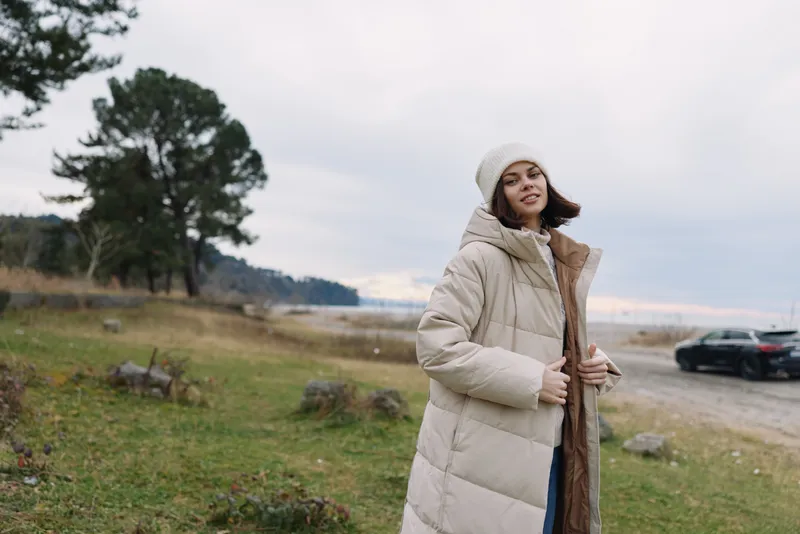 Winter fashion, woman in an oversized beige coat and knit hat posing outdoors in a grassy landscape with trees and a distant car, cloudy sky and calm mood for cold weather style