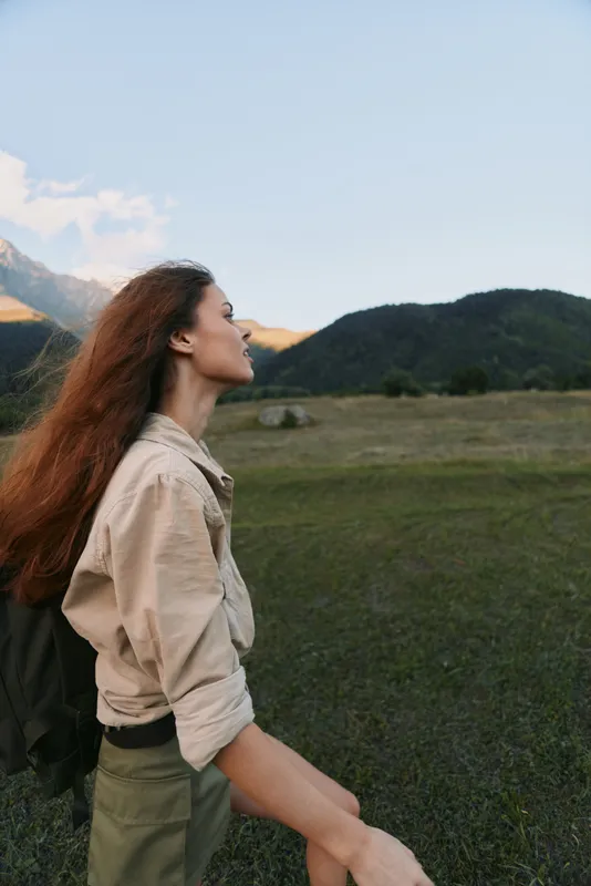 Young woman with long red hair enjoys a peaceful moment in a vast green meadow surrounded by mountains under a clear blue sky.