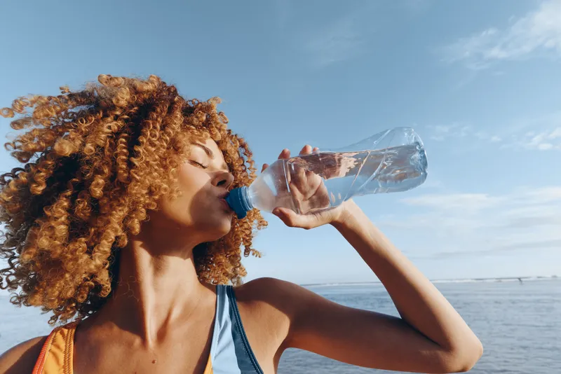 Woman with curly hair drinking water from a clear bottle by the sea under blue sky, hydration and wellness concept, outdoors summer refreshment after fitness, closeup side profile