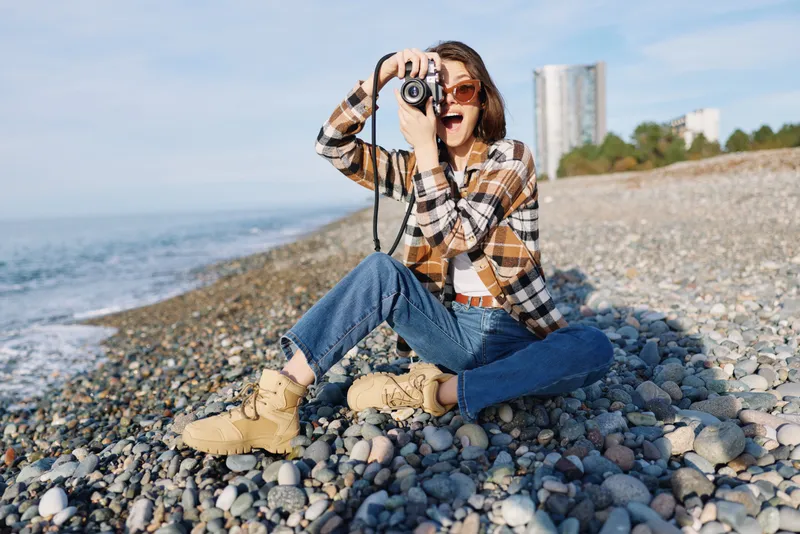 Female photographer on a pebble beach, wearing plaid shirt, jeans and boots, aiming a camera toward the sea under bright daylight for a casual outdoor shoot