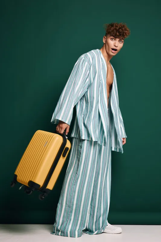 Young man in striped pajamas holding yellow suitcase standing against dark green background with surprised expression. Casual travel, luggage, fashion and lifestyle concept with studio lighting.