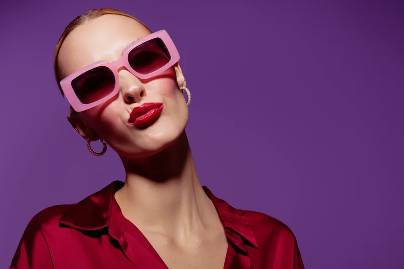 Stylish woman with pink sunglasses and red lipstick poses confidently against purple background. Fashion concept with bold colors and modern accessories.