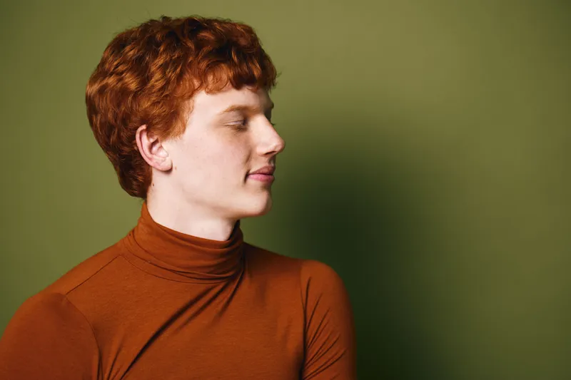 Portrait of a redhead woman with curly hair wearing brown turtleneck standing against olive green background with closed eyes and calm expression.