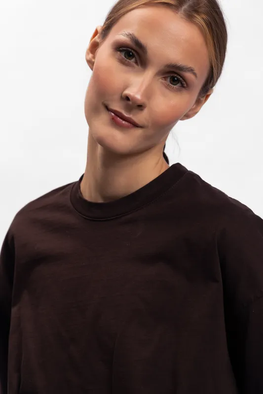 Young woman with light brown hair wearing a dark sweater poses against a white background. Soft natural expression with minimal makeup and casual style. Studio portrait.