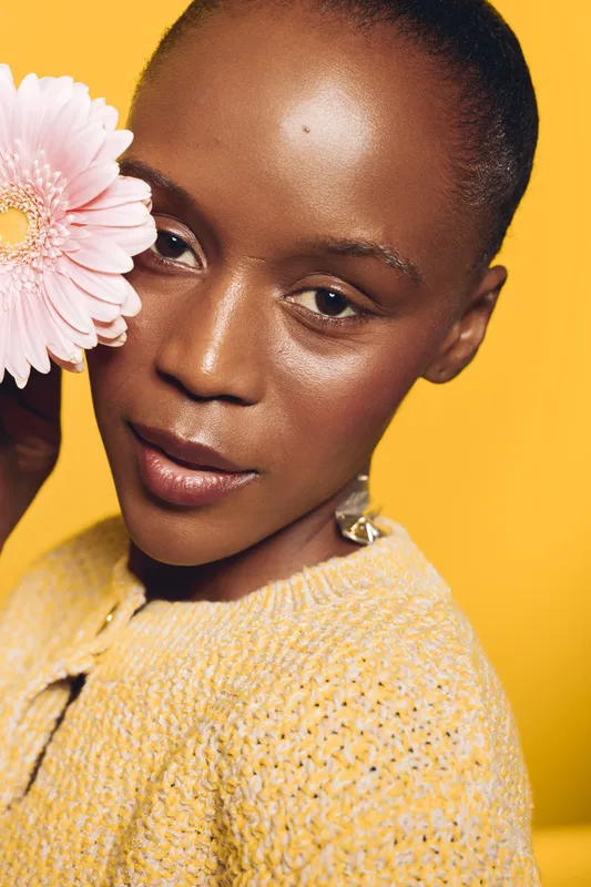 Close-up portrait of a dark skinned African woman with smooth skin holding a pink flower near her face. Yellow background and soft lighting create a warm, natural beauty concept.