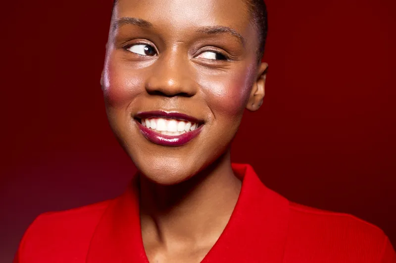 Portrait of a smiling dark skinned African woman wearing a red blazer with glossy lipstick and makeup, looking sideways on a red background. Concept of beauty, confidence, and fashion.
