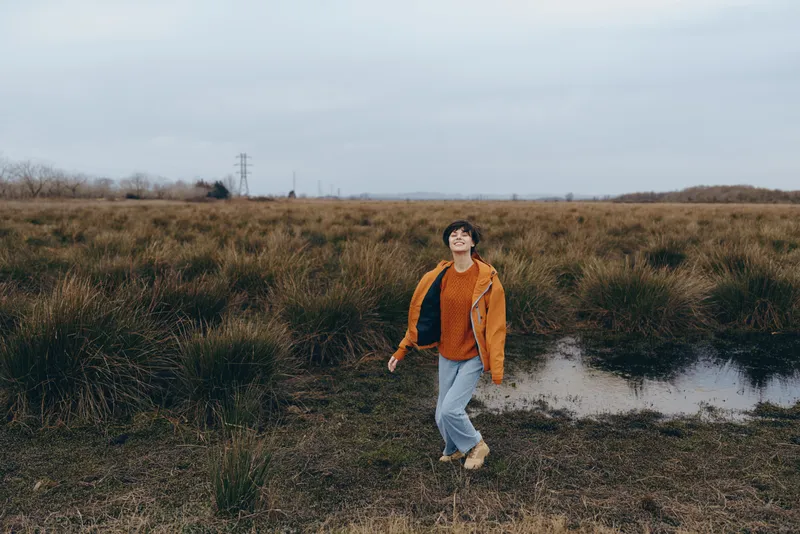 Woman walking outdoors in a grassy field by a pond, lifestyle nature landscape under cloudy sky, casual jeans and jacket, calm reflection near water, natural light, quiet horizon reeds