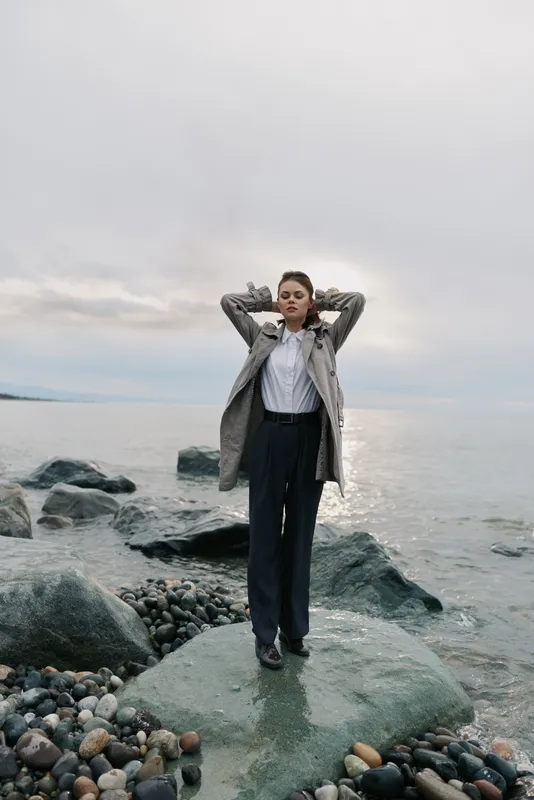Woman in elegant coat stands on rocky shore near calm sea with cloudy sky. She looks relaxed with hands behind head. Concept of nature, fashion, confidence and tranquility outdoors.