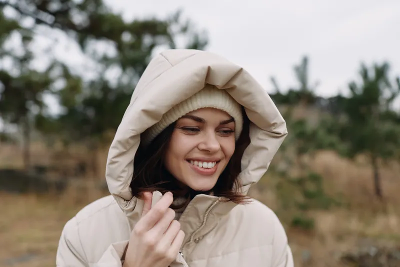 Woman in a beige hooded coat smiles outdoors, bundled for cold weather, portrait shot showing warmth, comfort and a calm natural landscape