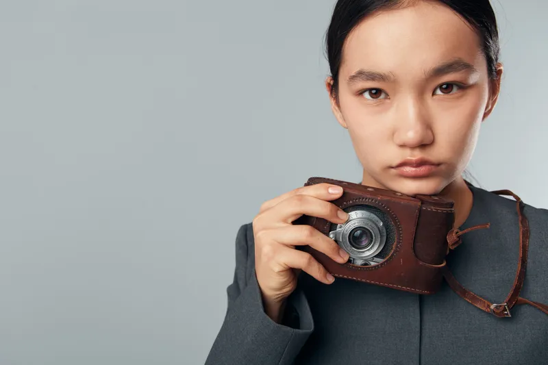 Young woman with vintage camera in hand looking at camera with serious expression on grey background. Portrait of female holding retro photo device closeup for hobby or photography.