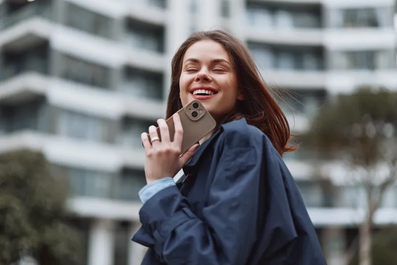 A smiling woman on a phone call outdoors in an urban setting beside a modern building, capturing a casual, confident moment for stock photography and storytelling.