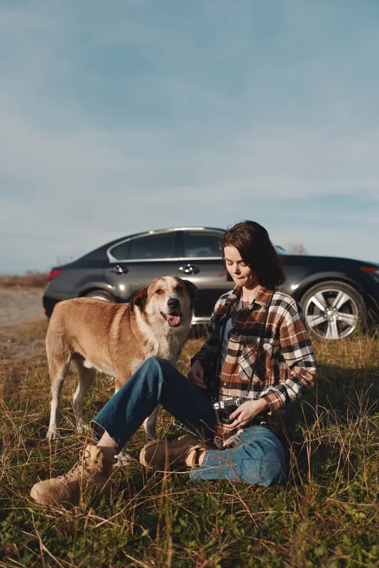 Dog and woman sit in a grassy field beside a parked car, enjoying a sunny day outdoors. Casual attire and a relaxed vibe capture companionship and nature during a roadside outing.