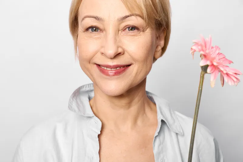Elderly woman smiling in a studio portrait, holding a pink flower on a clean white background, soft warm gentle expression, senior happiness wellbeing idea