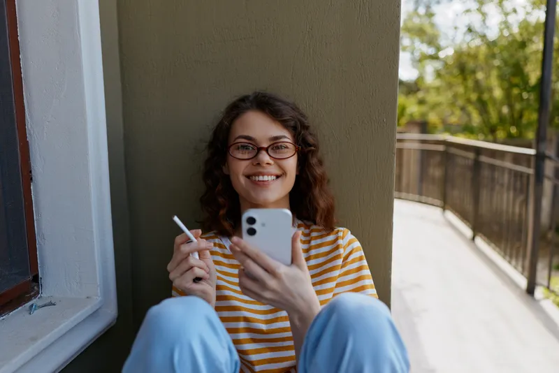 Smiling woman with glasses sits on a balcony, holding a cigarette in one hand and a smartphone in the other. Casual home lifestyle, relaxed mood, natural light, warm tones.