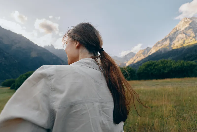 Woman in a light jacket stands in a meadow with distant mountains, capturing outdoor nature lifestyle, serene scenery, back view, sunlit day, peaceful mood.