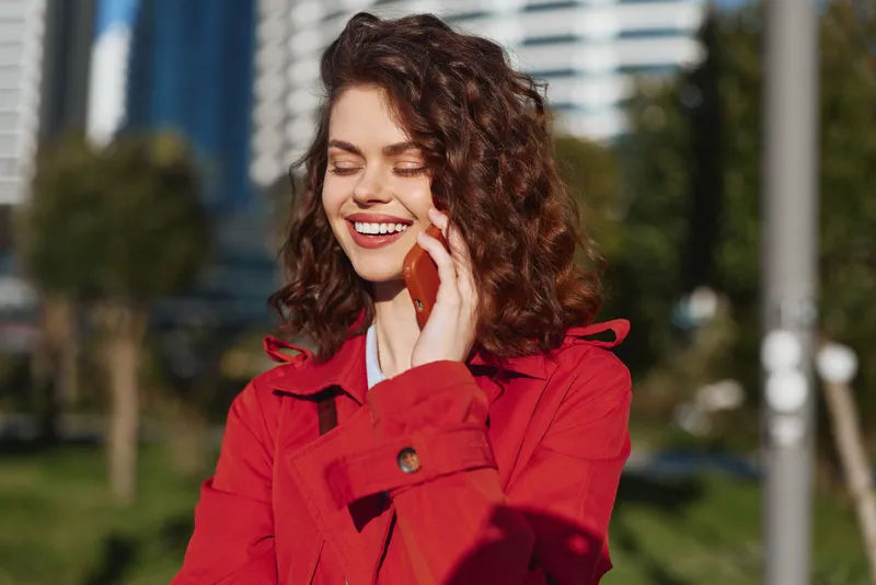 Smiling woman with curly hair wearing red coat talking on smartphone outdoors in urban park on sunny day. Concept of communication, happiness, and lifestyle.