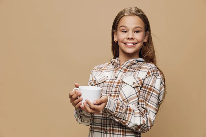 Girl smiling and holding white mug in both hands wearing plaid shirt against beige background. Portrait of child with long hair and happy expression in casual clothing.