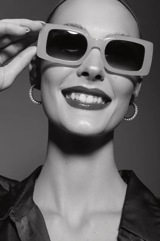 Smiling white woman with pale skin wearing oversized sunglasses and hoop earrings in studio. Concept of fashion, style, and beauty portrait.